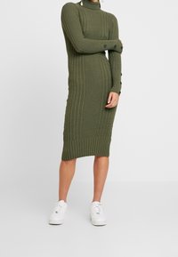 Mekko, joka on neulottu oliivinvihreästä langasta, jossa on ribbikuvio, korkea kaulus ja pitkät hihat, joissa on mustat napit. Yhdistetty valkoisiin tennareihin.