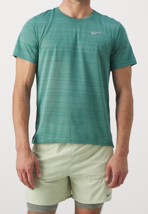 Homme portant un t-shirt de sport Nike à manches courtes couleur sarcelle et un short Nike vert clair avec un short de compression intégré, debout devant un fond uni.
