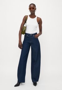 Jeans in denim scuro a gamba larga con vita alta e cuciture visibili. Abbinati a un top aderente bianco e una borsa verde.