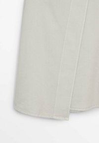 Gros plan de l'ourlet d'une jupe en tissu beige avec des détails de couture visibles et une texture subtile sur fond blanc uni.