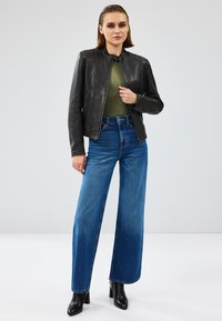 Giacca di pelle nera con colletto, abbinata a un top verde aderente e jeans blu a gamba larga. Stivali neri con tacco completano il look.