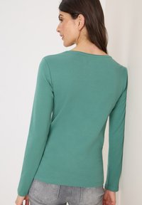 Groene blouse met lange mouwen van zacht materiaal. Heeft een brede halslijn en een rond hem. Het model draagt het met grijze jeans, gezien van achteren.