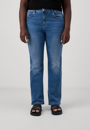 Person iført melletblå jeans med lige ben, sorte sandaler og en sort top, stående mod en ensfarvet lys baggrund.
