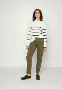 Sweater branco com riscas azul-marinho, combinado com calças de carga verde-azeitona e sapatos pretos tipo slip-on; modelo de pé contra um fundo simples.