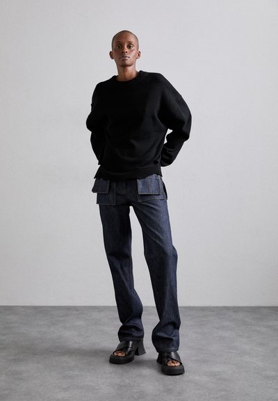 Pulover negru supradimensionat, pantaloni din denim bleu închis cu buzunare mari și sandale masive negre; textură netedă și design casual.