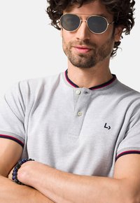 Jasnoszara koszulka polo z czerwonymi i niebieskimi akcentami, dwa guziki przy kołnierzyku i małe logo. Noszona z okrągłymi okularami przeciwsłonecznymi i bransoletką z koralików.