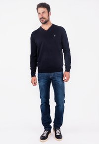 Pulover cu guler în V de culoare bleumarin, realizat dintr-un material moale, asortat cu blugi albaștri și adidași negri cu șireturi albe. Siluetă casual, ajustată.
