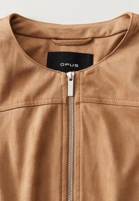 Giacca in suede beige con scollatura rotonda, caratterizzata da una chiusura frontale con cerniera e dettagli di cucitura minimalisti. L'etichetta riporta "OPUS."