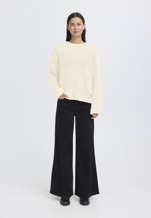 Maglione color crema, dal tessuto strutturato, con colletto rotondo e vestibilità ampia, abbinato a pantaloni neri a vita alta e gamba larga, accompagnati da calzature nere.
