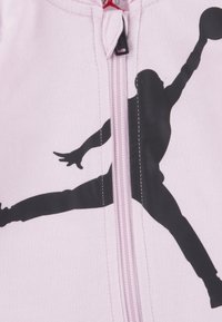 Sweat à zip rose clair présentant la silhouette noire d'un joueur de basketball en plein saut, avec une couture contrastante le long de la fermeture éclair.