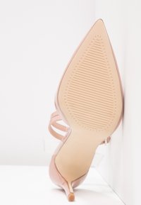 Chaussure à talon haut à bout pointu en rose nude avec semelle texturée et talon stiletto fin, posée en angle contre un fond blanc d'angle.