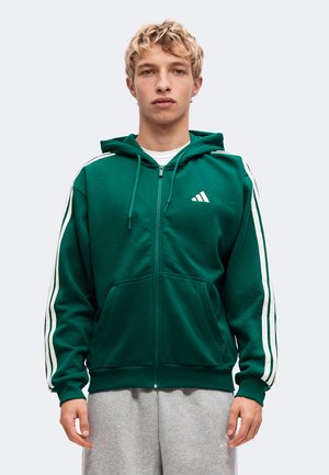 Ung mand iført en mørkegrøn lynlåshoodie med hvide striber på ærmerne og lysegrå joggingbukser, står mod en ensfarvet baggrund.