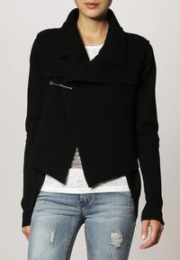 Svart stickad cardigan med sjalkrage, asymmetrisk front och en silverfärgad säkerhetsnål som accent. Mjukt material, slim fit och långa ärmar.