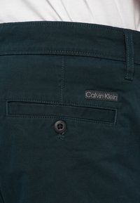 Tmavohnedé nohavice s zadným vreckom a čiernym gombíkom, na ktorom je štítok značky Calvin Klein, umiestnené na bielom látkovom pozadí.