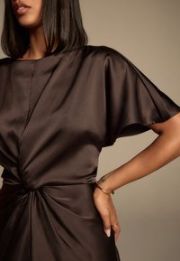 Brun tekstureret kjole med en afslappet pasform, der har en central vridningsdetalje ved taljen og flydende ærmer, hvilket skaber en glat silhuet.