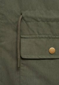 Olivově zelená látka s hladkým povrchem, vybavená horizontální kapsou, detaily z mosazného knoflíku a stahovací šňůrkou u otvoru.