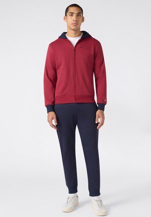Jeune homme portant un sweat à capuche rouge zippé avec des poignets et une capuche bleu marine, un pantalon de jogging bleu marine et des baskets blanches, debout devant un fond uni.