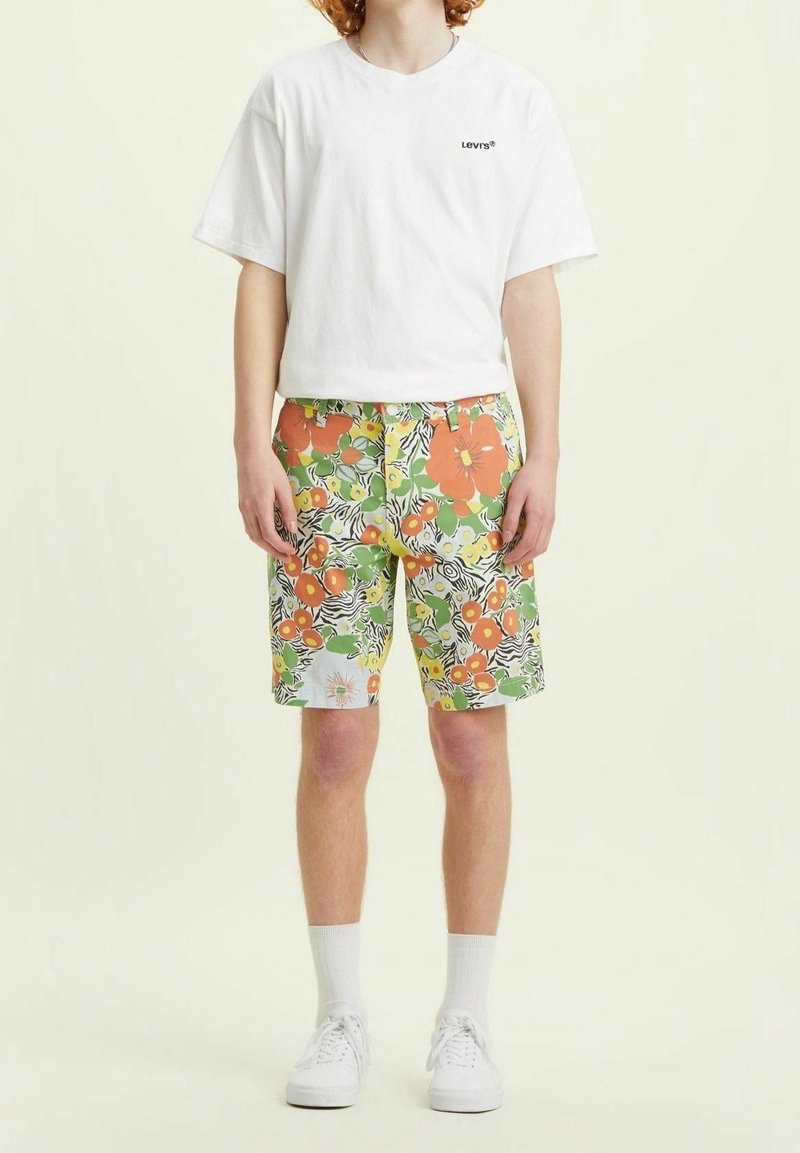 Personne portant un t-shirt Levi's blanc, un short coloré à motifs floraux et feuillus, des chaussettes blanches et des baskets blanches, debout devant un fond clair.