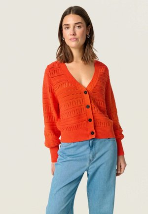 Oranje gebreide cardigan met een V-hals, voorzien van een textuurpatroon, lange mouwen en vijf zwarte knopen aan de voorkant. Gecombineerd met blauwe jeans.