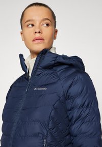 Marineblå pufferjakke med hette, med glidelås foran og sidelommer. Strukturert, quiltet design med Columbia-logo på brystet.