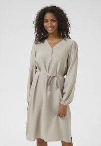 Robes beige à manches longues, patte de boutonnage et détail de taille nouée. Tissu texturé avec des poignets élastiques et des fentes sur les côtés pour la mobilité.
