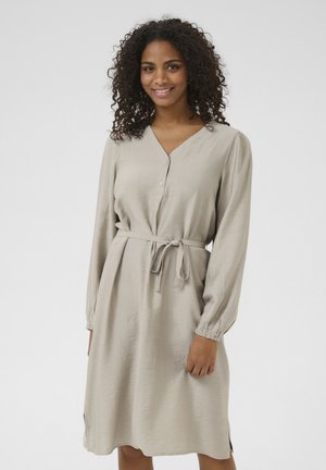 Robes beige à manches longues, patte de boutonnage et détail de taille nouée. Tissu texturé avec des poignets élastiques et des fentes sur les côtés pour la mobilité.