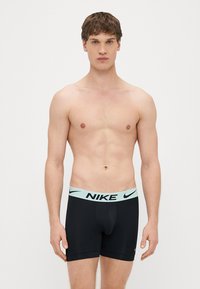 Mustad bokserid helesinise Nike logoga vöökohal, keha järgi lõigatud ja sileda tekstuuriga, kantud palja ülakehaga mehemodellil.