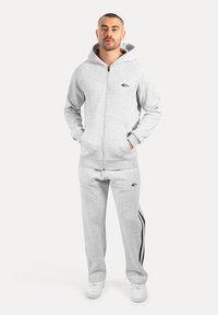 Sudadera gris con cremallera y cordón, con bolsillos tipo canguro, combinada con pantalones de chándal grises a juego que tienen rayas laterales negras, usados con zapatillas blancas.