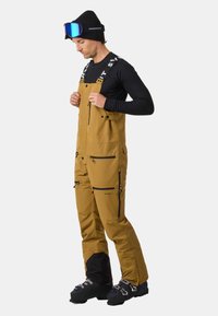 Skrócony kombinezon narciarski w brązowym materiale z czarnymi akcentami. Wyposażony w regulowane szelki, liczne kieszenie na zamek oraz wzmocnione dolne nogawki.