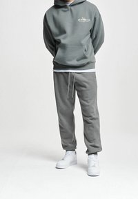 Personne portant un sweat à capuche gris avec poche avant, un pantalon de survêtement gris assorti et des baskets blanches, debout les mains dans le dos sur fond blanc.