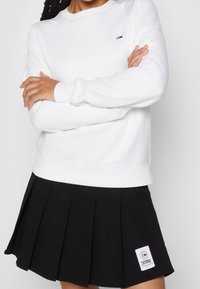 Sweatshirt bianco con collo rotondo e polsini a coste, abbinato a una gonna plissettata nera con patch del logo. Tessuto liscio e vestibilità classica.