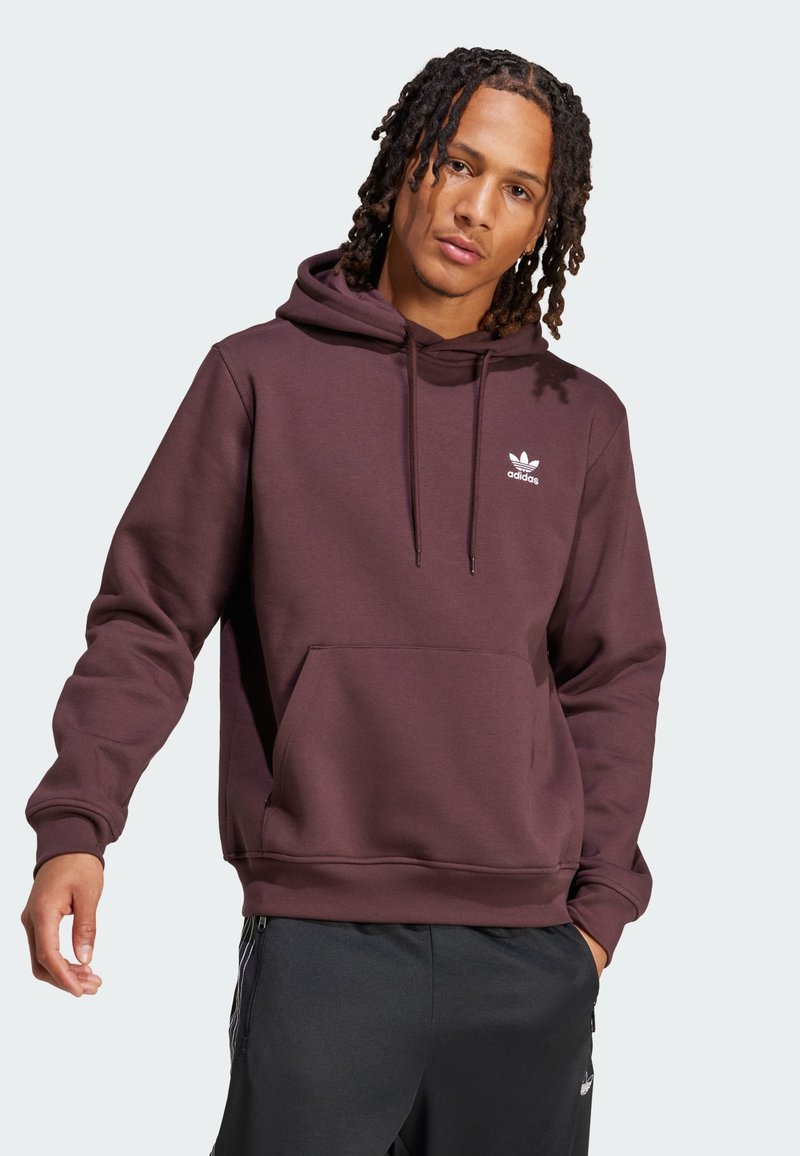 Brun hettegenser med frontlomme, med hvit Adidas-logo på brystet. Løs passform med ribbestrikkede mansjetter og nederkant.