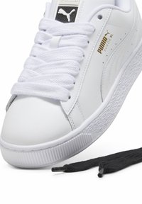Baskets blanches Puma en daim taille XL avec des lacets blancs noués, accompagnées d'une paire de lacets noirs détachés sur fond blanc.