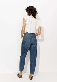 Blouse blanche avec des manches volantées et des détails en œillets, portée avec un jean bleu taille haute. Le modèle porte des ballerines imprimées léopard.