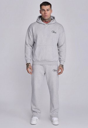 Mężczyzna z tatuażami na szyi i ręce, ubrany w jasnoszary kaptur i pasujące spodnie dresowe, stojący na jednolitym tle.