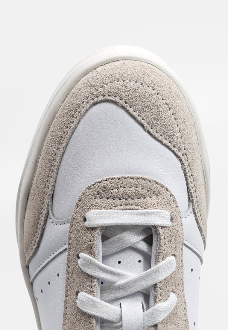 Zapatilla blanca de cuero con acentos en ante gris claro, puntera redonda, detalles perforados y cordones planos blancos. Se destacan las texturas contrastantes.
