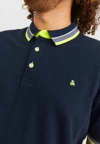 Marineblauwe poloshirt met een contrasterende neongroene kraag en afwerking, voorzien van drie knoopsluitingen en een klein geborduurd logo.