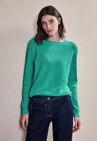 Zelený žebrovaný svetr s kulatým výstřihem a diagonálním texturovaným vzorem, kombinovaný s tmavými džíny. Měkká tkanina a dlouhé rukávy viditelné.