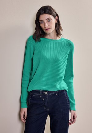 Pull vert côtelé avec un col rond et un motif texturé diagonal, associé à un jean foncé. Tissu doux et manches longues visibles.