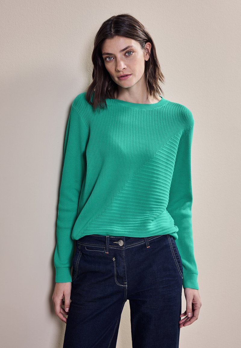 Zelený žebrovaný svetr s kulatým výstřihem a diagonálním texturovaným vzorem, kombinovaný s tmavými džíny. Měkká tkanina a dlouhé rukávy viditelné.