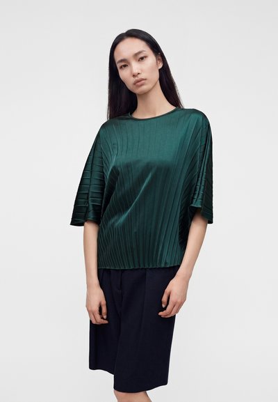 Femme aux longs cheveux foncés portant un haut vert plissé à manches courtes et un short noir au genou, debout devant un fond uni.