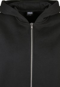 Sweat à capuche zippé noir avec une fermeture éclair argentée, capuche avec cordon et une étiquette "Urban Classics" taille XS dans le col.