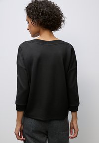 Sweat-shirt noir avec un col large, des manches trois-quarts et des accents texturés aux épaules. Porté avec un pantalon gris à motifs.