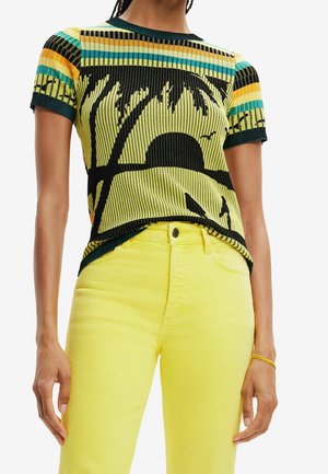 Persona que lleva una camisa de manga corta con un diseño de puesta de sol y palmera y pantalones amarillos brillantes de talle alto, de pie con los brazos relajados.