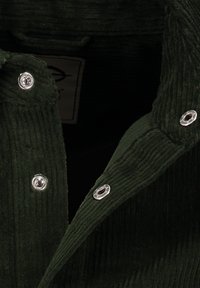 Grüne Cordjacke mit ausgeprägter Rippstruktur, ausgestattet mit silbernen Druckknöpfen und einem umgeschlagenen Kragen. Das Innere zeigt das Etikett.