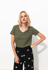 T-shirt vert olive à col en v en tissu doux associé à un pantalon noir taille haute avec des motifs brodés blancs.