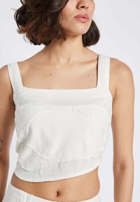 Weißes, kurzes Tanktop mit quadratischem Ausschnitt, texturierten Details und geschichteten Stoffakzenten. Hergestellt aus einem weichen Material, das den Bauch zeigt.