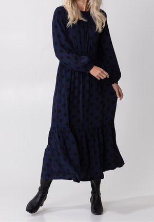 Kobieta ubrana w długą granatową sukienkę w czarne kropki z długimi rękawami, w czarnych butach sięgających do połowy łydki, stojąca na jednolitym tle.