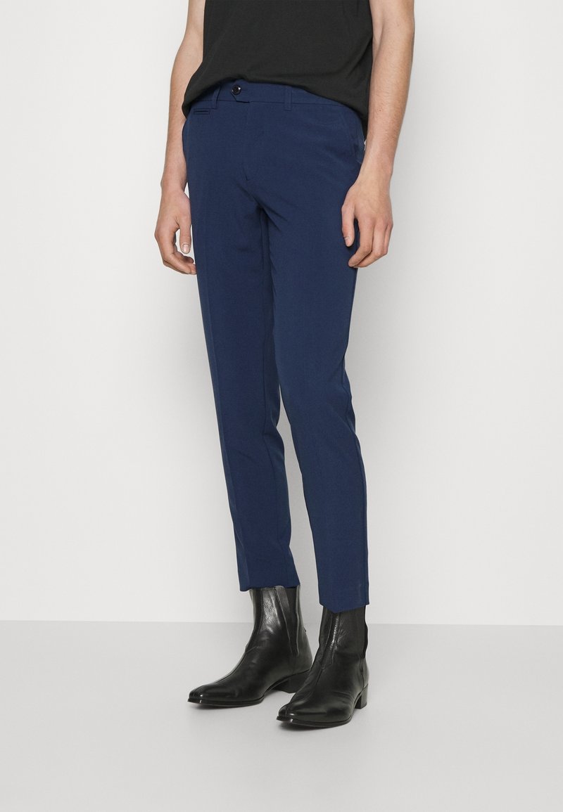 Pantalon bleu marine slim associé à des bottines en cuir noir, porté par une personne vêtue d'un haut noir sur un fond uni.