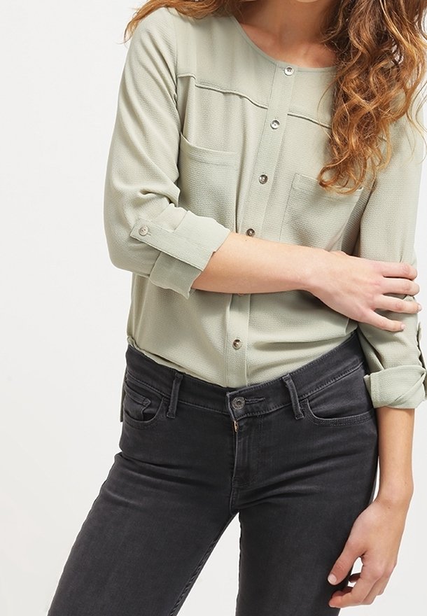Femme portant un chemisier vert clair boutonné avec les manches retroussées et un jean gris foncé, debout avec un bras croisé sur le torse.